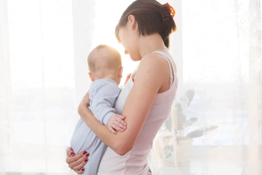 A young nanny stands with her baby in the bedroom against a window.jpeg